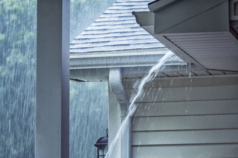 Rain storm water is gushing and splashing off the tile shingle roof - pouring over the overhanging eaves trough aluminum roof gutter system on a suburban residential colonial style house near Rochester, New York State, USA during a torrential July mid summer downpour.